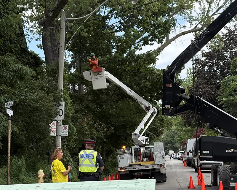 Utility crew working from a bucket truck beside a residential street, with traffic cones and a police officer directing traffic